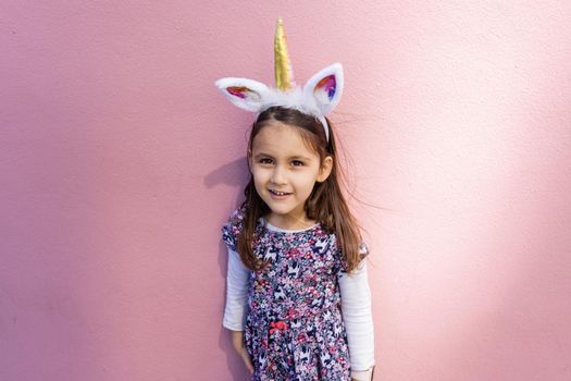 Adorable view of happy little girl wearing unicorn headband with pink background. Portrait of cute smiling child with unicorn horn and ears in front bright pink wall. Lovely kids in costumes