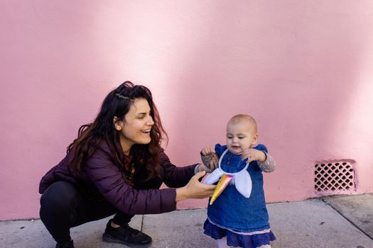 Portrait of happy mother in squatting position holding baby with unicorn headband in front of pink wall. Lovely view of smiling woman next to cute baby girl in blue dress. Happy family outdoors