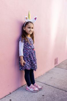 Adorable view of happy little girl wearing unicorn headband with pink wall as background. Portrait of cute smiling child with unicorn horn and ears standing on the sidewalk. Lovely kids in costumes