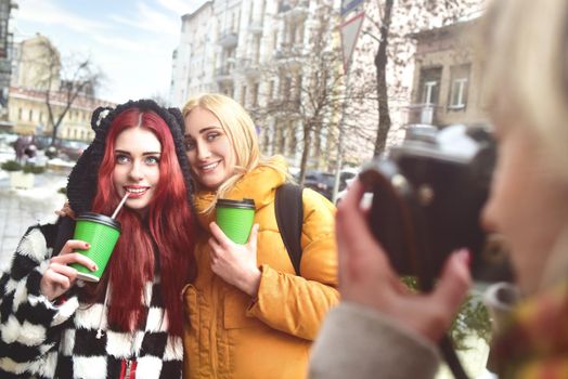Two cute girlfriends hold their coffee and pose for the camera while her friend takes their picture.