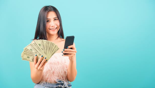 Smiling face portrait asian beautiful woman her usring mobile smart phone and holding bunch money looking at camera on blue background, with copy space for text