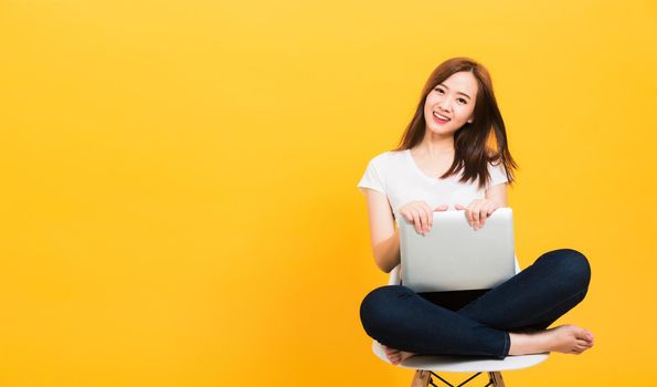Asian happy portrait beautiful cute young woman teen smiling sitting crossed legs on a chair with laptop computer looking to camera isolated, studio shot on yellow background with copy space