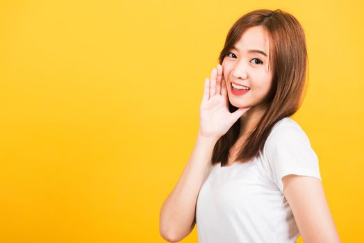 Asian happy portrait beautiful cute young woman teen standing wear t-shirt hand on mouth talking whispering secret rumor looking to camera isolated, studio shot on yellow background with copy space