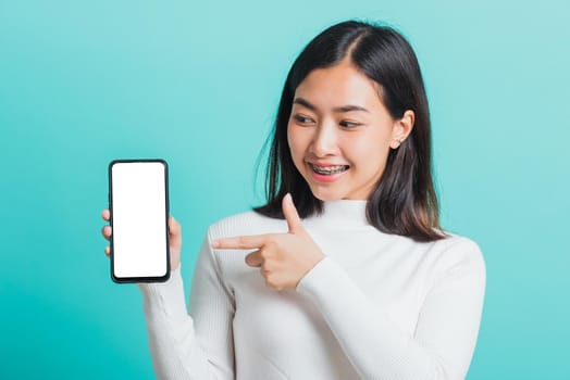 Beautiful Asian woman smile holding a smartphone on hand and pointing finger to the blank screen, female excited cheerful her show mobile phone isolated on a blue background, Technology concept