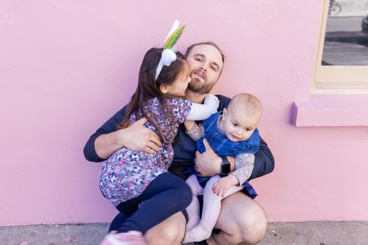 Portrait of little girl lovely hugging her father while holding cute baby in front of a pink wall. Adorable view of smiling bearded man sitting and holding joyful daughters. Happy family outdoors