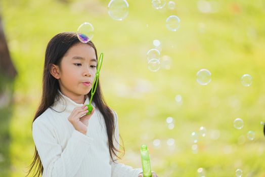 Happy Asian little cute girl child having fun and enjoying outdoor play blowing soap bubbles during in the garden park on a sunny day, summer time