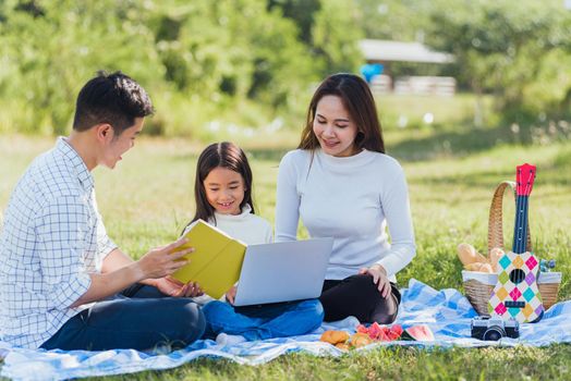 Happy Asian lifestyle young family father, mother and little girl having fun outdoor sitting on picnic blanket using laptop computer technologies while relaxing to open song at garden park in summer