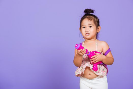 Thai child funny hold toy water pistol and smile, Happy Asian little girl holding plastic water gun, studio shot isolated on purple background, Thailand Songkran festival day national culture concept