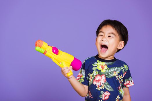 Happy Asian little boy holding plastic water gun, Thai kid funny hold toy water pistol and smiling, studio shot isolated on purple background, Thailand Songkran festival day national culture party