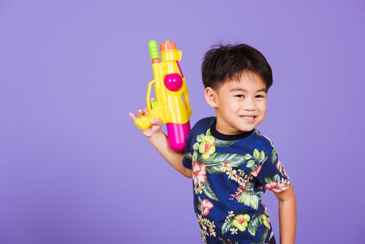 Thai kid funny hold toy water pistol and smiling, Happy Asian little boy holding plastic water gun, studio shot isolated on purple background, Thailand Songkran festival day national culture concept