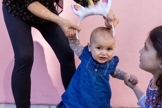 Beautiful view of woman and little girl holding cute baby with unicorn headband. Adorable baby girl holding her mother and older sister with pink wall as background. Happy family portraits