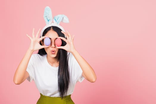 Happy beautiful young woman smiling wearing rabbit ears holding colorful Easter eggs front eyes, Thai female with bunny ear hold easter egg covering eye, studio shot isolated on pink background