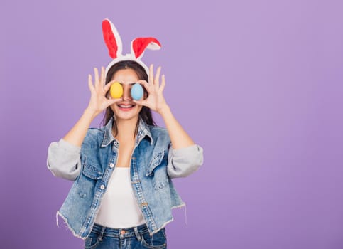 Happy beautiful young woman smiling wearing rabbit ears and denims holding colorful Easter eggs front eyes, Thai female with bunny ear, easter egg cover eye, studio shot isolated on purple background