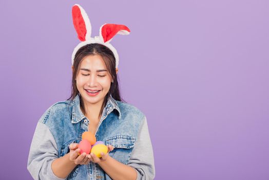 Happy Easter concept. Beautiful young woman smiling wearing rabbit ears and denims hold colorful Easter eggs gift on hands, Portrait female looking at egg, studio shot isolated on purple background