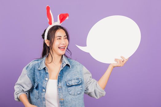 Happy Easter Day. Beautiful young woman smiling excited wearing rabbit ears and denims holding empty speech bubble, Portrait female looking at bubbles, studio shot isolated on purple background