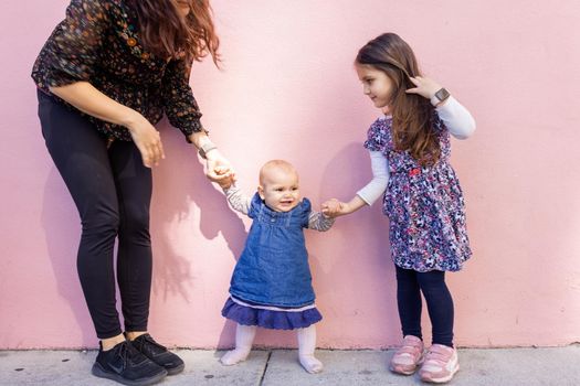 Portrait of woman and little girl holding cute smiling baby by the hands with pink wall as background. Adorable baby standing on sidewalk between her mother and older sister. Happy family outdoors
