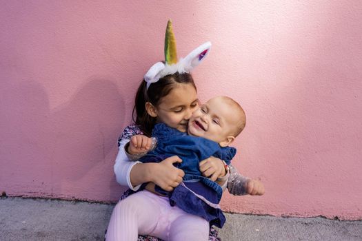 Adorable view of little girl wearing unicorn headband hugging and kissing her baby sister with pink background. Portrait of cute children sitting in front bright pink wall. Lovely kids in costumes