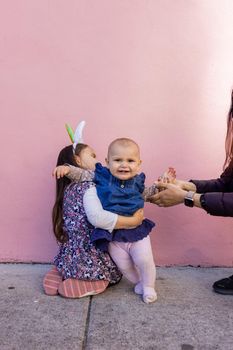 Adorable view of little girl wearing unicorn headband hugging her happy baby sister with pink background. Mother holding cute toddler hand while sitting in front bright pink wall. Lovely kids outdoors