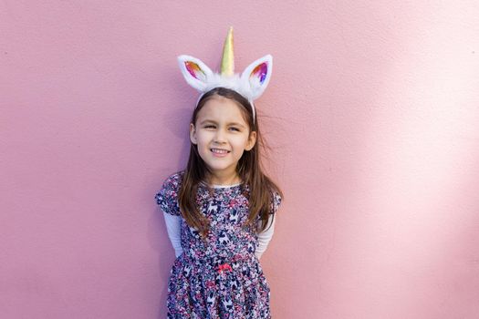 Adorable view of happy little girl wearing unicorn headband with pink background. Portrait of cute smiling child with unicorn horn and ears in front bright pink wall. Lovely kids in costumes