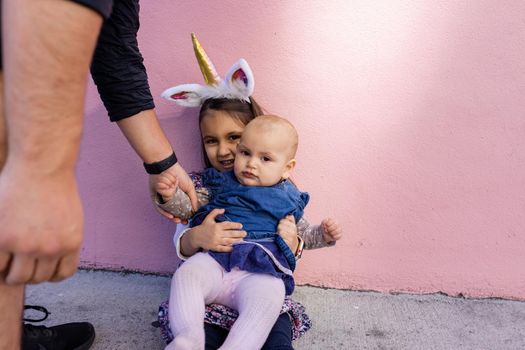 Adorable view of little girl wearing unicorn headband kissing her baby sister with pink background. Father holding cute toddler hand while sitting in front bright pink wall. Lovely kids in costumes