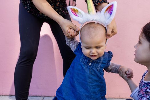 Beautiful view of woman and little girl holding cute baby with unicorn headband. Adorable baby girl holding her mother and older sister with pink wall as background. Happy family portraits