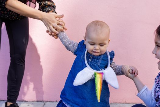 Beautiful view of woman and little girl holding cute baby with unicorn headband. Adorable baby girl holding her mother and older sister with pink wall as background. Happy family portraits