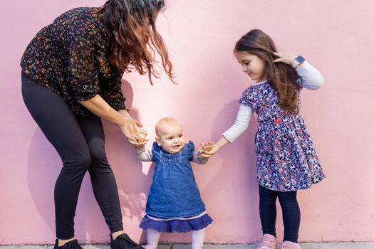 Portrait of woman and little girl holding cute smiling baby by the hands with pink wall as background. Adorable baby standing on sidewalk between her mother and older sister. Happy family outdoors