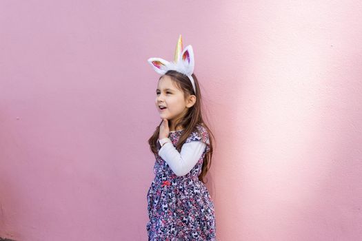 Adorable view of happy little girl wearing unicorn headband with pink background. Portrait of cute smiling child with unicorn horn and ears in front bright pink wall. Lovely kids in costumes