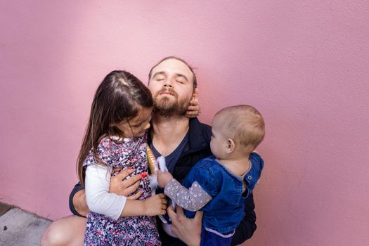 Portrait of peaceful father hugging his adorable baby and older daughter in front of pink wall. Sunlight on bearded man with eyes closed while holding baby and little girl. Happy family outdoors