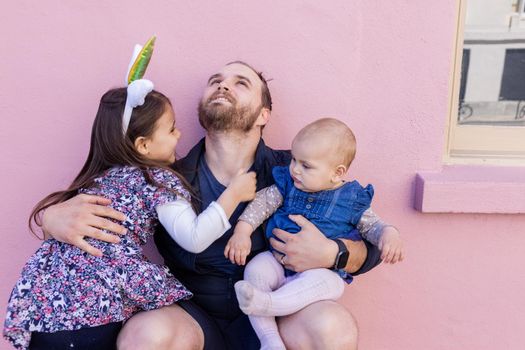 Portrait of young father looking up and hugging his adorable baby and older daughter in front of a pink wall. Bearded man in squatting position holding baby and little girl. Happy family outdoors