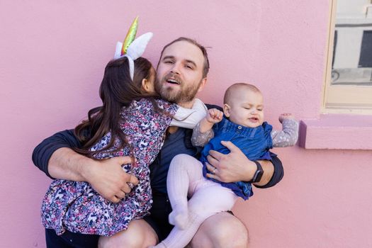 Portrait of adorable little girl lovely hugging her father while holding cute baby in front of a pink wall. Smiling bearded man holding joyful and cute daughters. Joyful family outdoors