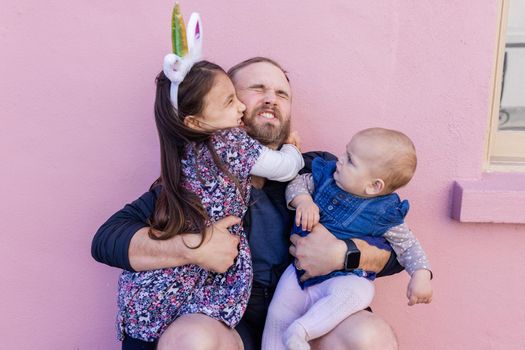 Portrait of adorable little girl lovely hugging her father while holding cute baby in front of a pink wall. Bearded man with struggling facial expression holding cute daughters. Happy family outdoors