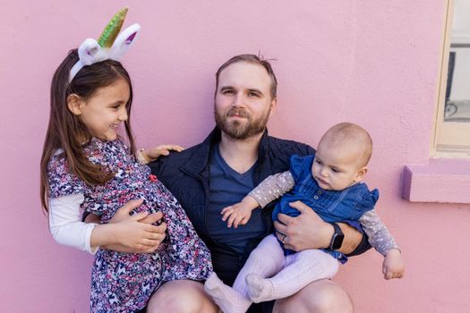 Portrait of young father hugging his adorable baby and older daughter in front of a pink wall. Bearded man sitting and holding joyful cute little girl and baby. Happy family outdoors