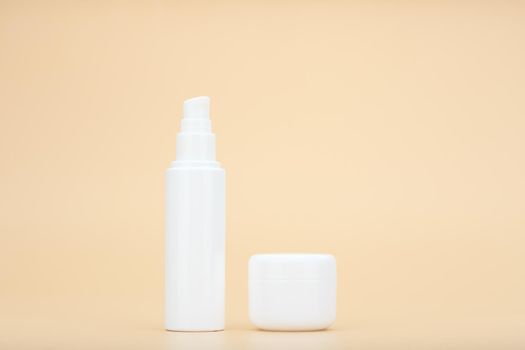 Minimalistic still life with unbranded white tube with beauty product, face cream, serum or lotion and white jar with mask or scrub for daily skin care routine against beige background with copy space