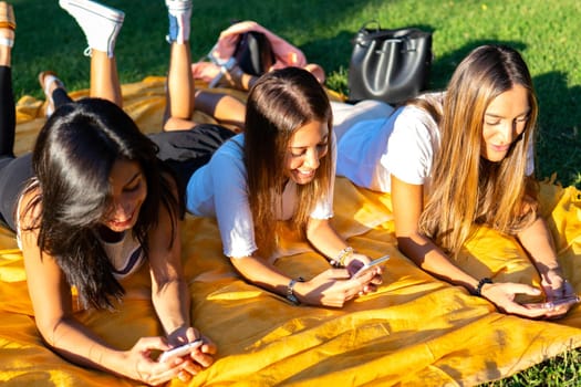 Three young best beautiful friends spending time in social network together lying on grass meadow in city park at sunset or dawn. Girls having fun with smartphone using internet mobile technology