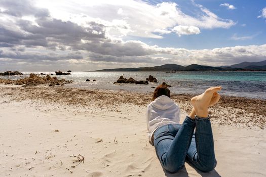 Lonely girl lying on the sand at the beach in winter ocean sea with dramatic blue sky full of clouds. Pensive woman in casual jeans clothes look at the horizon with barefoot in nature contemplation