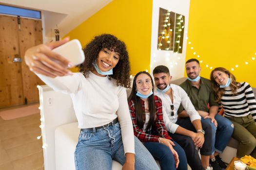 Group of young people at home wearing lowered protective medical mask posing for selfie sitting on a couch at home party, Afro american girl using smartphone to share on social network happy moments