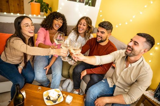 Team of young happy people celebrating at home toasting with champagne wine glasses sitting at the sofa in the living room. Millennial student drinking together in holiday party
