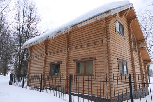 A large log house behind the fence in winter against the snow and a roof in the snow, icicles hang from the roof. High quality photo