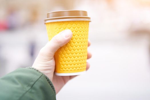 unrecognizable man holding yellow paper cup with coffee in hand on blured street background