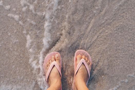 Woman feet wear slippers stand on sand tropical beach with sea water splash background.