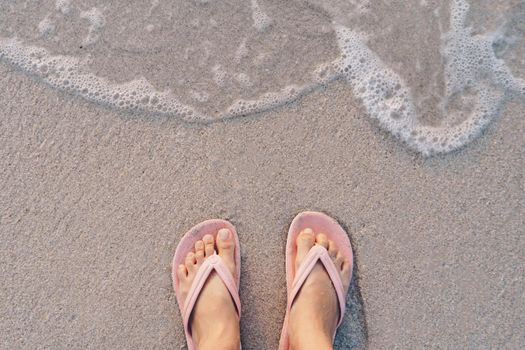 Woman feet wear slippers stand on sand tropical beach with sea water splash background.