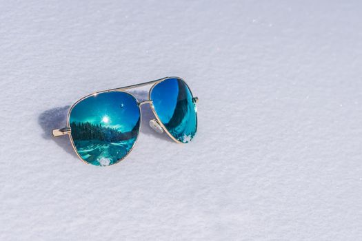 Sunglasses with blue lenses in which the sun and the forest are reflected lie on the snow