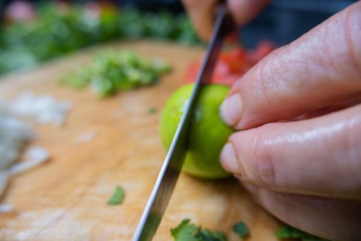 Close-up of hands slicing lime on wooden cutting board with chopped tomatoes, onions, and peppers. Person cutting fresh vegetables into pieces on wood surface. Traditional sauce preparation