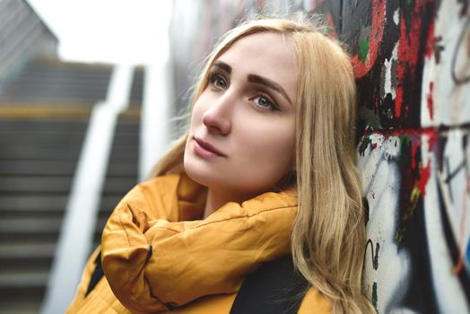 Portrait of a beautiful, pretty, attractive teenage blonde with a pensive look leaning back against a graffiti-painted wall outdoors, close-up