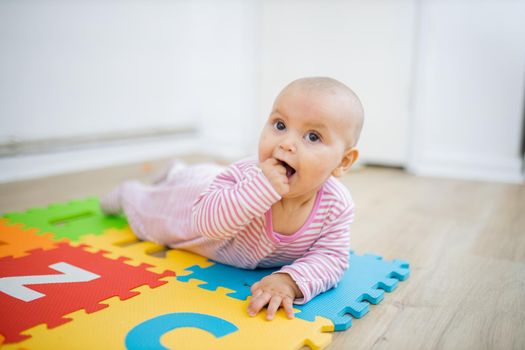 Adorable baby lying face down on colorful children mat with her fist in her mouth. Portrait of curious baby playing on the floor. Happy babies having fun