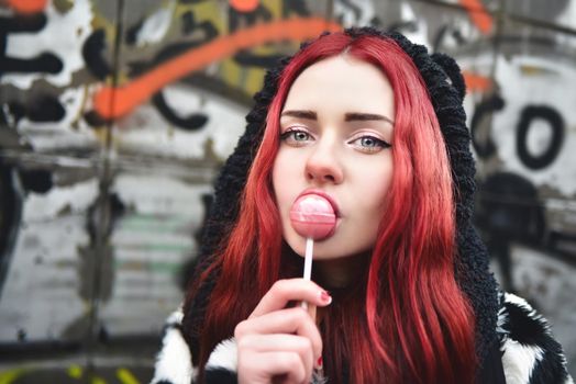 Portrait of a young beauty in a hoodie with a delicious pink lollipop, posing against a wall of graffiti. Positive Emotions. Urrban style