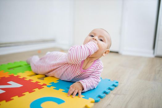 Adorable baby lying face down on colorful children mat with her fist in her mouth. Portrait of curious baby playing on the floor. Happy babies having fun