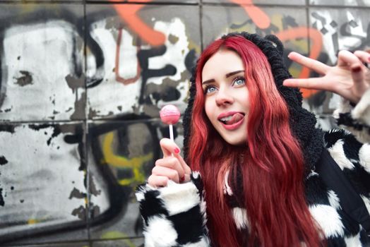 Portrait of a young beauty teen girl in a hoodie with a delicious pink lollipop, showing peace sign into the camera. Positive Emotions. Urrban style