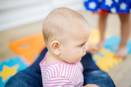 Adorable baby siting on colorful foam mat between the feet of her father. Portrait of cute baby sitting on the floor. Happy babies having fun